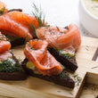 Load image into Gallery viewer, Crostini topped with seasoned cream cheese and smoked salmon slices on a wooden cutting board.

