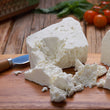 Load image into Gallery viewer, Block of fresh cheese with a knife on a wooden board, surrounded by tomatoes and herbs.

