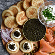 Load image into Gallery viewer, Overhead caviar spread on a round wooden board with blini, crème fraîche with chives, smoked salmon with dill, red onion, capers, and an open tin of dark green ossetra caviar.

