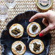 Load image into Gallery viewer, Hand reaching for blinis topped with crème fraîche and caviar on a dark marble plate with Petrusco tin and vodka shots.
